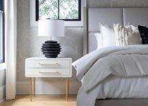 Bedroom features a light gray wood and fabric bed atop a black and white rug and a black lamp that illuminates a white and gold nighstand on white and gray wallpaper.