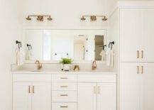 Off white washstand with brass hardware and white quartz featuring brushed gold faucets under a white wooden framed full-length vanity mirror illuminated by 3-light brass sconces.