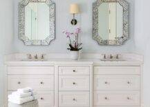 Two antiqued French mirrors hang on either side of a Camille Long Sconce over a white dual bath vanity adorned with polished nickel knobs and polished nickel faucets. A white rope stool sits on ivory mosaic floor tiles in front of the vanity.