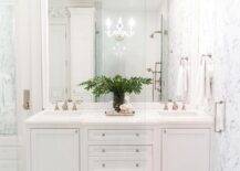 A white beveled mirror is hung from a white and black wallpapered wall over a white dual bath vanity accented with glass knobs and a polished marble countertop. The washstand sits on white, gray, and black hexagon floor tiles.