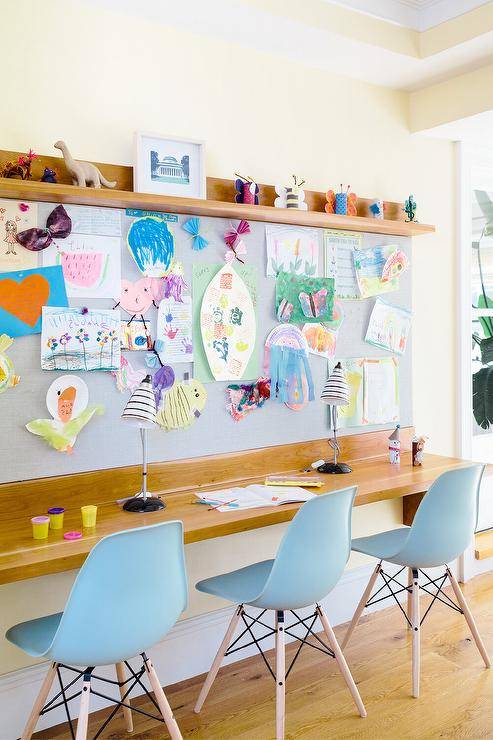 Powder blue molded plastic chairs are placed at a wood top wall mount desk fixed beneath a blue linen pin board hung under a wood shelf.