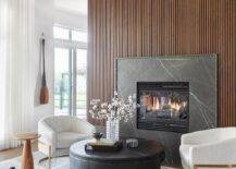 Living room features a brown reeded wall with black marble fireplace mantle, modern white and gold chairs with a round gold and black coffee table atop a gray rug and a small round accent table.