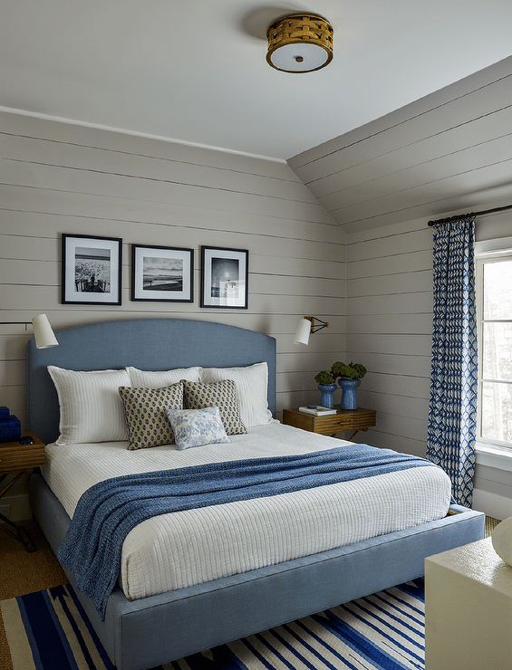 Blue and gray bedroom design features a blue upholstered bed with blue and white bedding flanked by X base bedside tables with blue flower vases lit by brass sconces on gray shiplap trim and a blue stripe rug with blue curtains.