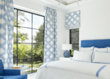 White and blue bedroom boasts a blue upholstered bed dressed in white and blue border bedding paired with a white nightstand and blue lamp.