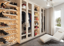 Pink-beige, l-shaped closet shoe shelves are mounted in a corner beside glass front wardrobe cabinets flanking light bag shelves. A white boucle lounge chair sits on a gray rug facing the wardrobes.