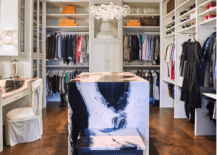 Spacious custom walk in closet features an island with a black and white marble waterfall edge and white built in shelves.