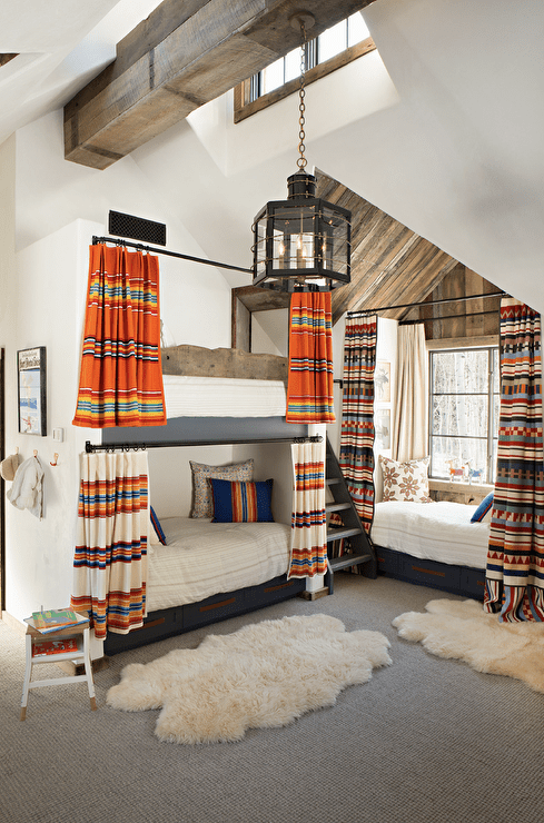 Beautifully styled cabin bunk room features a rustic built-in bunk bed complemented with striped curtains and drawers donning brown leather pulls. Sheepskin rugs sits sit on gray carpeting in front of the bunk bed and in front of a daybed hidden beneath a window and behind red, blue, and orange curtains.