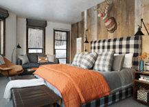 Welcoming country bedroom boasts a gorgeous black and white gingham bed complemented with an orange quilt and black plaid pillows. The bed sits on a gray rug against a wall covered in rustic wood vertical panels. A decorative animal bust is mounted above the headboard, while the bed is lit by oil rubbed bronze swing arm sconces fixed over wooden nightstand with metal grill accents.