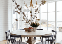 A brass chandelier hangs over a round, brown dining table paired iwth black spindle dining chairs. The dining space is positioned on a stained wood floor in front of windows framed by white shiplap.